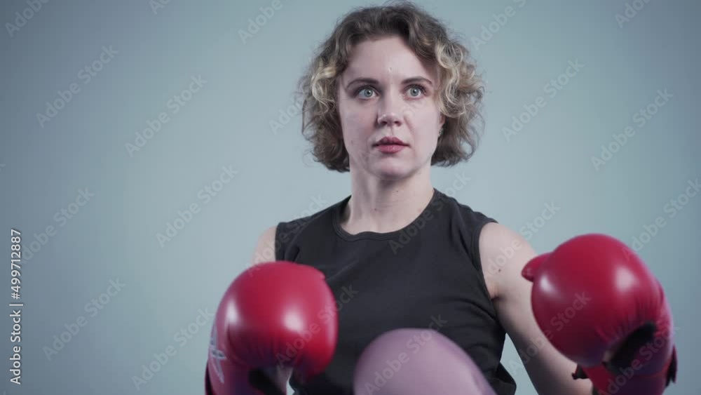 Young beautiful slim woman posing in red boxing gloves in gym against the background of a gray wall during a photo shoot, backstage video. fitness woman in red boxing gloves posing in fighting stance