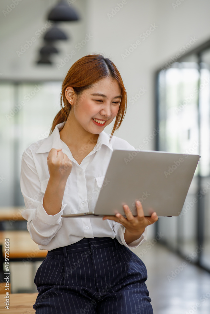 Image of a happy excited Asian young woman using a laptop computer and looking at the screen, isolated over blue wall background making a winner gesture. Image of a happy excited Asian young woman using a laptop computer and looking at the screen, isolated over blue wall background making a winner gesture.