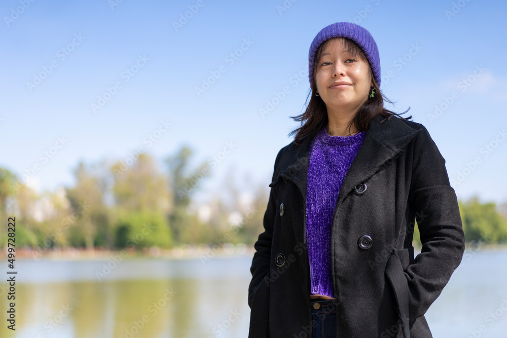 Portrait of a young Latin woman dressed in a pullover, coat and wool cap, looking at the camera, at a lake with copy space
