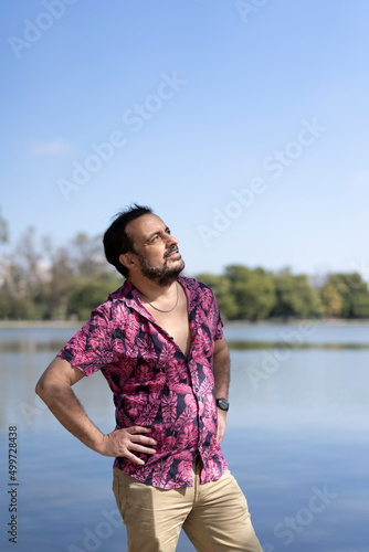 Bearded mature man wearing a fuchsia shirt, arms akimbo, looking at the camera at a lake