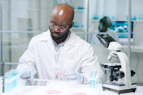 African American male scientist sitting by workplace in laboratory and scrolling in tablet while searching for online data