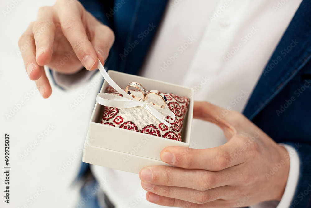 The groom holds gold wedding rings in a decorative box with a small pillow and ribbons for the bride in the morning indoors on a white background on the wedding day The groom holds gold wedding rings in a decorative box with a small pillow and ribbons for the bride in the morning indoors on a white background on the wedding day