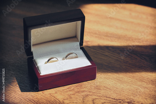 Gold wedding rings accessory for the bride and groom in a red decorative box inside light on a table with a wooden coating in yellow tone © Ihor
