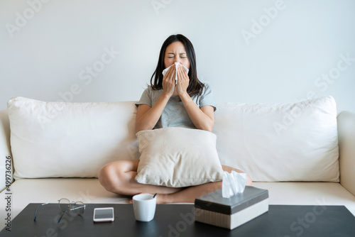 Photos Sick young Asian woman sitting on couch blowing her nose on a tissue conceptual of healthcare , seasonal flu, rhinitis or an allergic reaction in hay fever