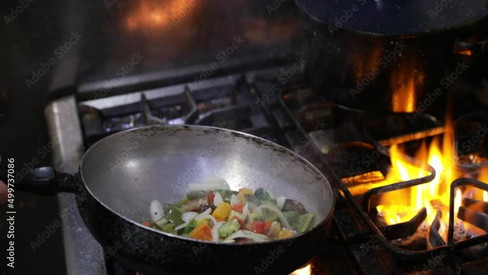 Culinary. Asian cuisine. Closeup view of a chef cooking sliced vegetables in the wok pan. The food catches fire and the chef shakes the wok. Culinary. Asian cuisine. Closeup view of a chef cooking sliced vegetables in the wok pan. The food catches fire and the chef shakes the wok.