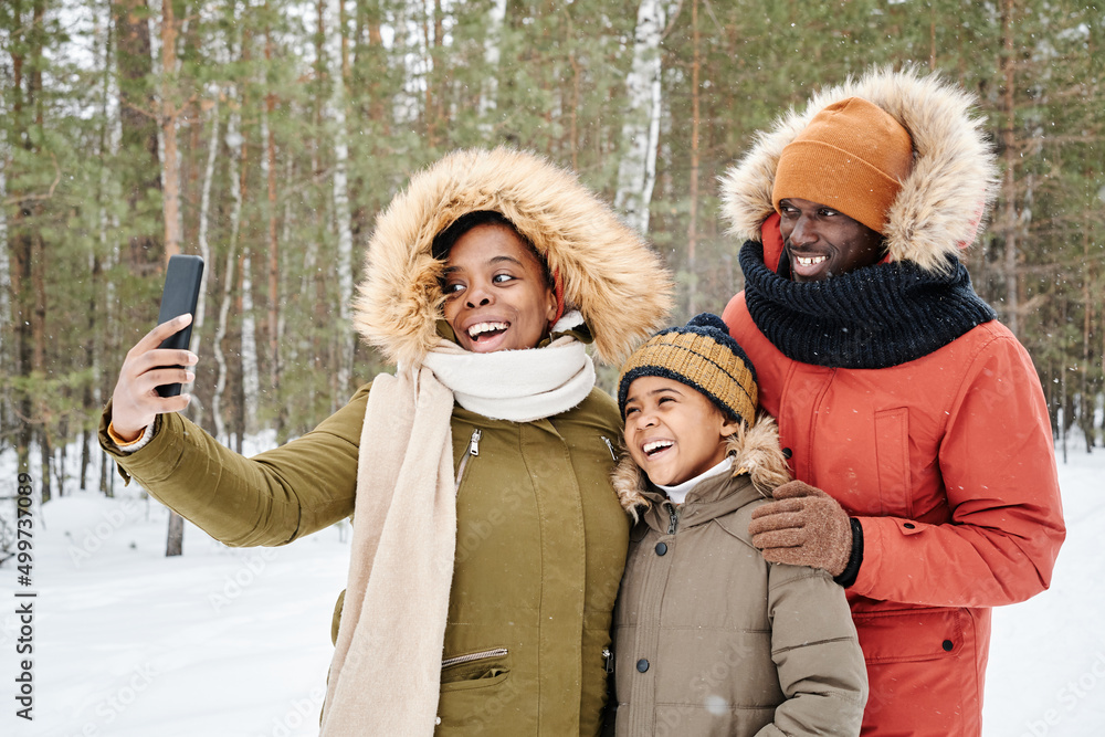 Happy affectionate African American family in warm winterwear making selfie in park or forest while woman holding smartphone