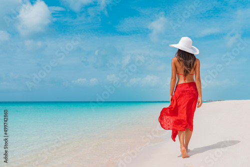 Luxury beach vacation elegant tourist woman walking relaxing in red beachwear and sunhat on white sand Caribbean beach. Lady tourist on holiday vacation resort