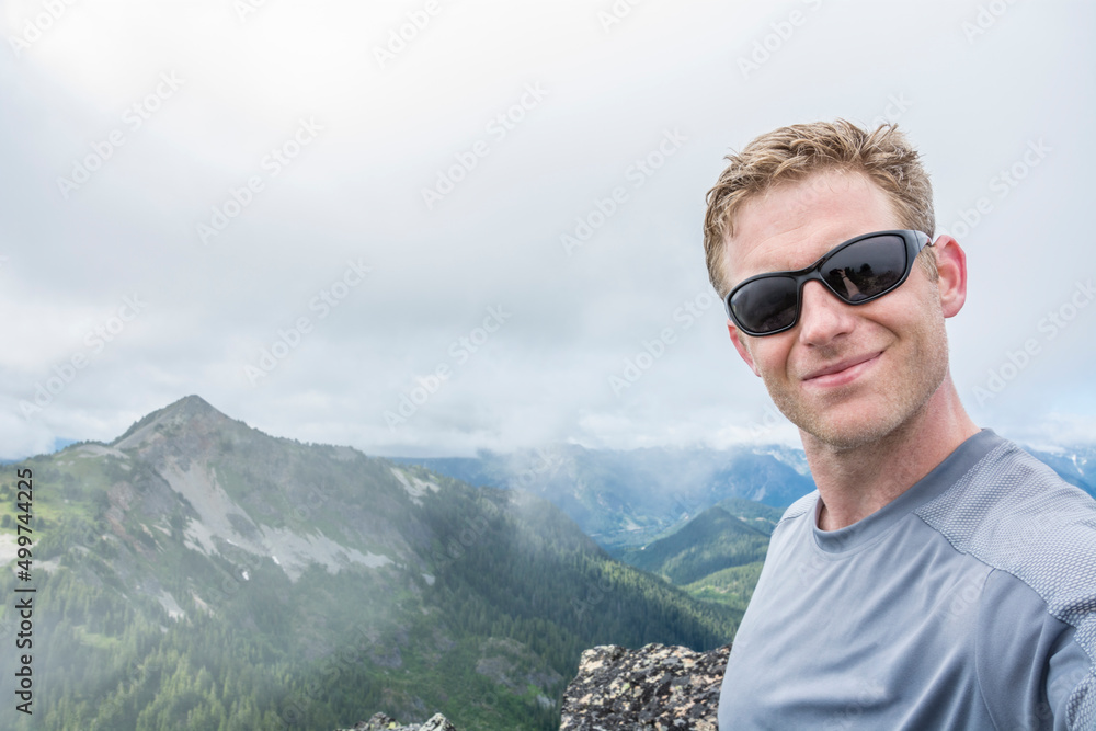 Adventurous athletic male hiker standing on top of a mountain taking a selfie while wearing sunglasses on a cloudy day. 