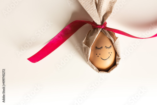 Easter egg with a painted mouth and eyelashes with napkin rabbit ears on a white background. Easter Ukraine 2022. Cool egg for Easter with a painted face and a red ribbon © Dubnytskaya Photo