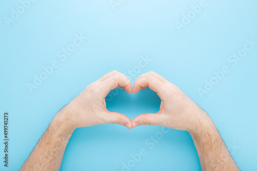 Heart shape created from young adult man hands on light blue table background. Pastel color. Love and happiness concept. Closeup. Point of view shot. Top down view.