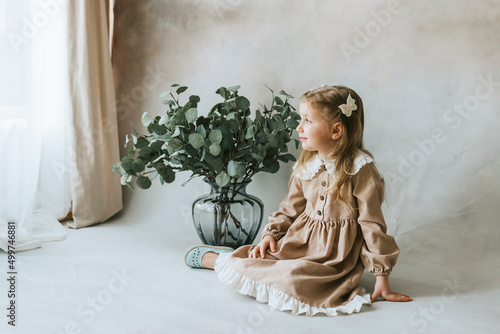 little blonde girl in vintage dress near retro background and vase with green branches looks thoughtfully out window on spring day, happy childhood, simple and cozy life © klavdiyav