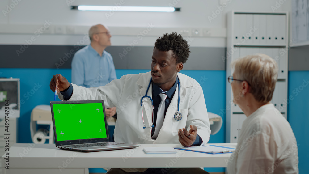 Young medic having laptop with green screen technology on desk while talking to elder patient at healthcare clinic. Horizontal device with isolated mockup template and chroma key background Young medic having laptop with green screen technology on desk while talking to elder patient at healthcare clinic. Horizontal device with isolated mockup template and chroma key background