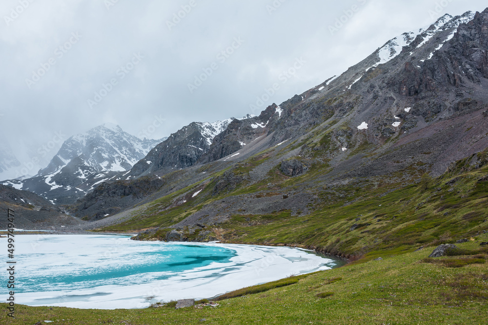 Obraz premium Atmospheric mountain landscape with frozen alpine lake and high snowy mountains. Awesome overcast scenery with icy mountain lake on background of snow mountains in low clouds. Scenic view to ice lake.
