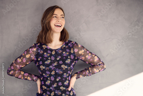 Portrait of young happy woman, looking beautiful with a big smile on her mouth. Posing with hands on the waist and looking right, standing isolated against concrete background in a beautiful dress.	