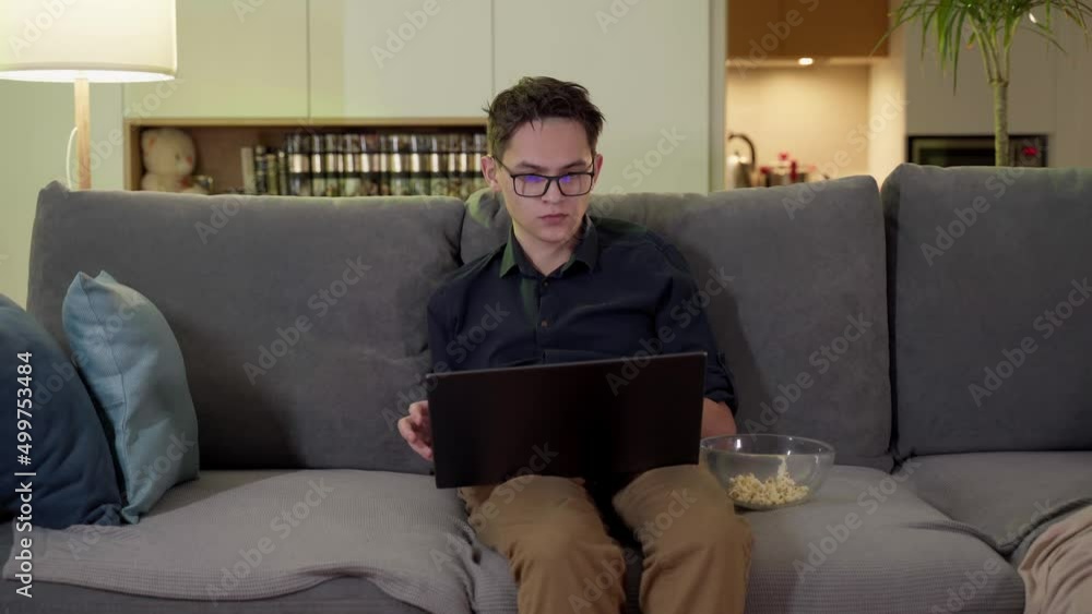 A young guy with glasses sits on a sofa with a laptop. Remote work, online. A young guy with glasses sits on a sofa with a laptop. Remote work, online.