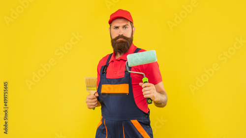 Bild auf Leinwand bearded man painter in work clothes hold paint roller and brush on yellow backgr