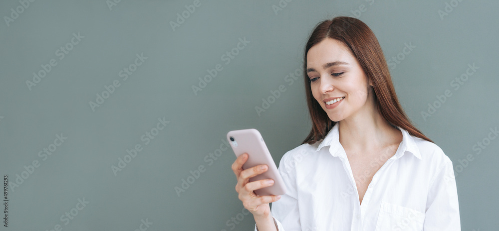 Young smiling woman student with dark long hair in white shirt using mobile phone on grey background, banner Young smiling woman student with dark long hair in white shirt using mobile phone on grey background, banner