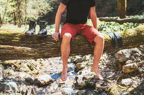  Man sitting on the fallen tree log over the mountain forest stream while he waiting for socks laundry drying and trekking boots. Active people traveling, hiking or trekking concept image.