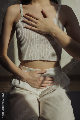 Crop woman in white clothes touching chest and stomach while breathing during yoga meditation at home in room with bright sunlight 