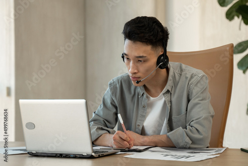 Asian assistant writes down payment details on paper helping customer to make purchase. Young man works using headset and laptop at table in office