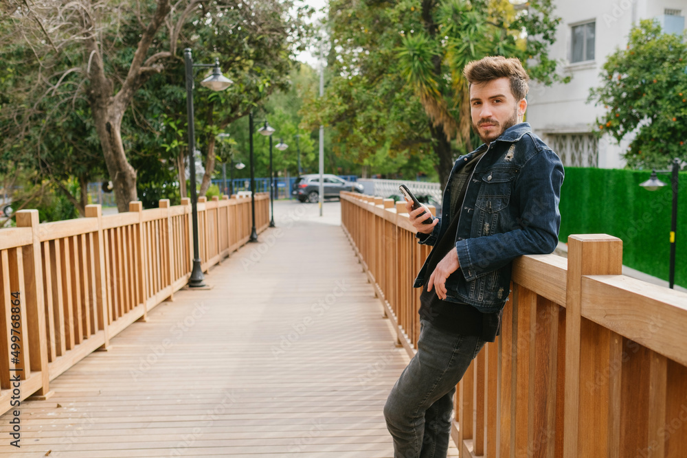 Fototapeta premium Posing, the young bearded handsome man is wearing a denim jacket, She leans her back on the bridge and poses for the camera, has a serious facial expression