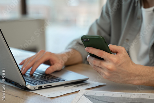 Young man assistant types personal details to clients on laptop holding mobile phone in hand. Professional worker sits at wooden table in office closeup