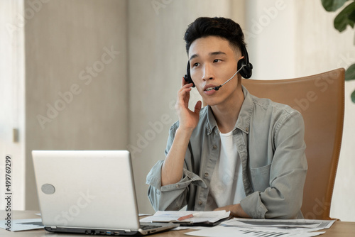 Asian assistant writes down payment details on paper helping customer to make purchase. Young man works using headset and laptop at table in office