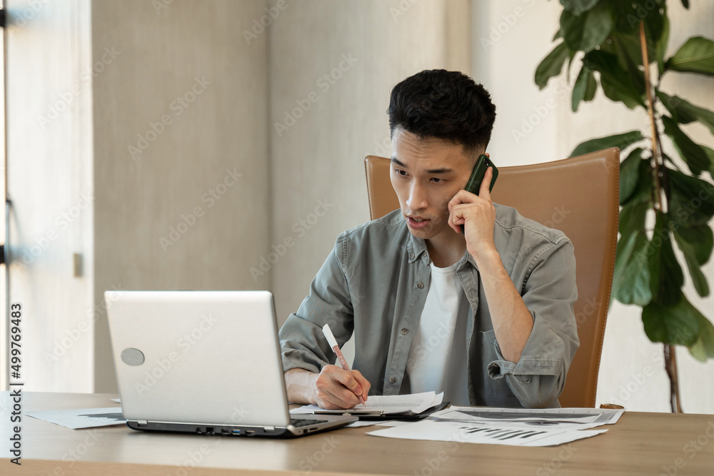Young Asian male secretary calls to manager explaining difficult issue at work by mobile phone. Busy employee sits near laptop at wooden table in office Young Asian male secretary calls to manager explaining difficult issue at work by mobile phone. Busy employee sits near laptop at wooden table in office