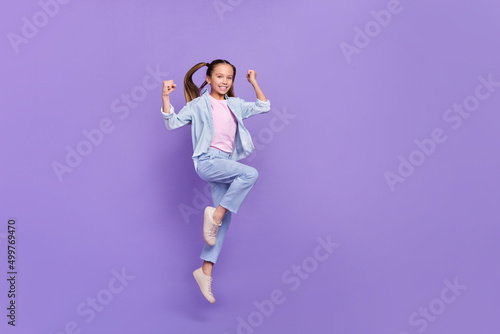 Photo of excited pretty little school girl wear denim shirt rising fists jumping high empty space isolated purple color background