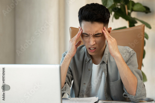 Young Asian male employee holds head trying to solve difficult issue at work in office. Desperate man sits near laptop at table feeling unsure closeup