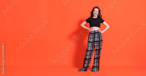 Young brunette girl in black and white trouser suit stands in full growth on orange background, looks into camera and smiles. © mityru