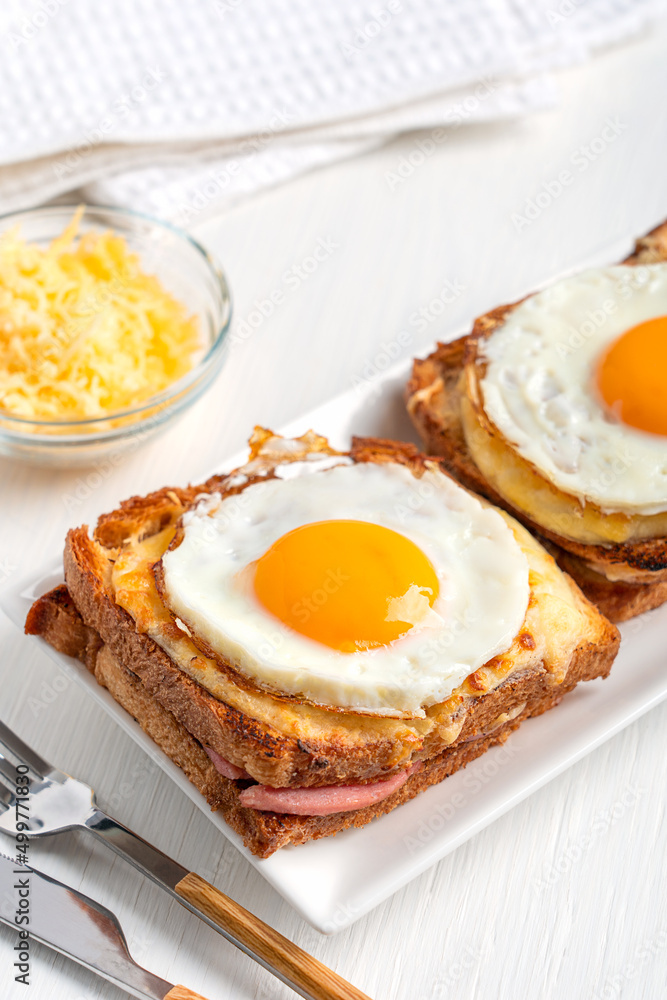 Homemade Croque Madame French hot sandwiches made of ham and cheese inside toasted bread topped with fried egg with yolk served for breakfast on plate with tableware and towel on white wooden table Homemade Croque Madame French hot sandwiches made of ham and cheese inside toasted bread topped with fried egg with yolk served for breakfast on plate with tableware and towel on white wooden table