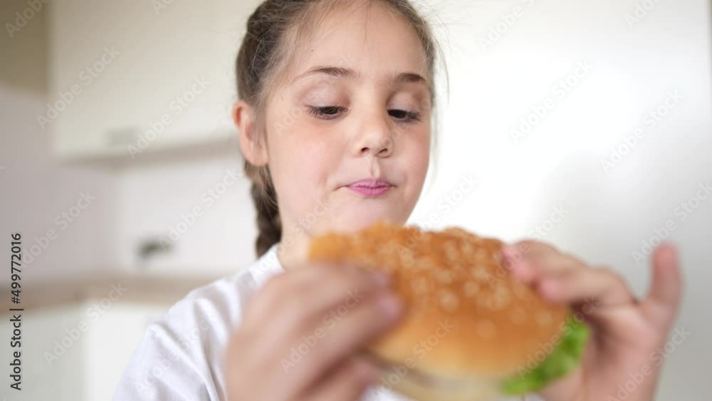 little girl eating a hamburger. unhealthy fast food proper nutrition concept. child greedily with pleasure bites a big burger in the kitchen at home. kid eats meal fast food close-up little girl eating a hamburger. unhealthy fast food proper nutrition concept. child greedily with pleasure bites a big burger in the kitchen at home. kid eats meal fast food close-up