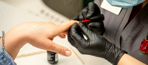 Manicure varnish painting. Close-up of a manicure master wearing rubber black gloves applying red varnish on a female fingernail in the beauty salon