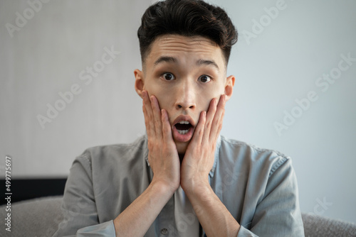 Young Asian man gets scared watching horror TV program against white wall. Terrified guy puts hands on cheeks and opens mouth sitting on sofa closeup