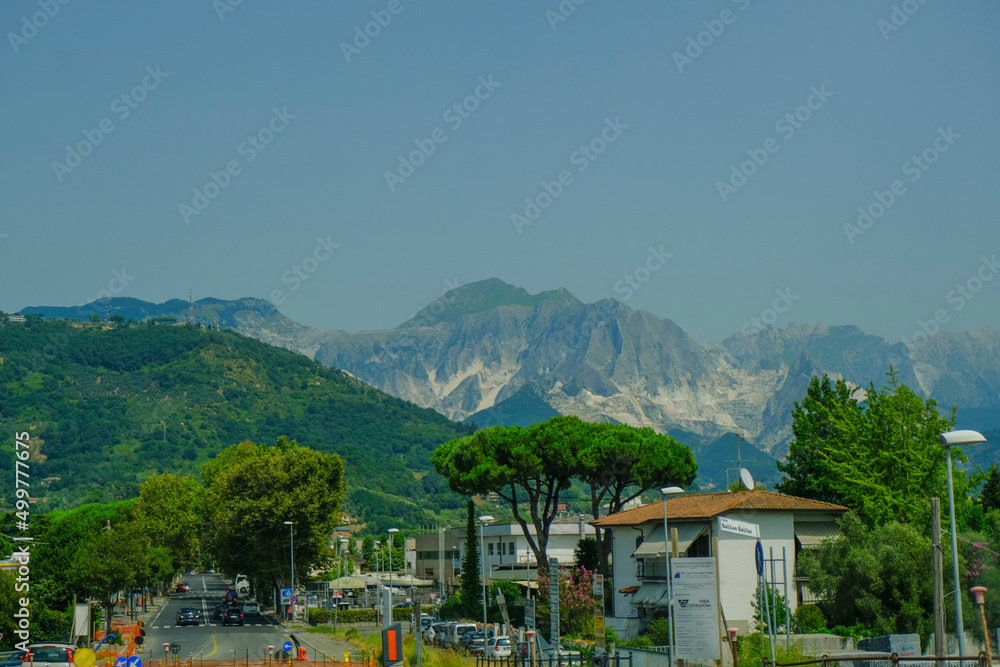 Fototapeta premium Backdrop of the Apuane mountains in Tuscany, Marina di Carrara, Italy. Mountains landscape with pines, city streets across blue sky