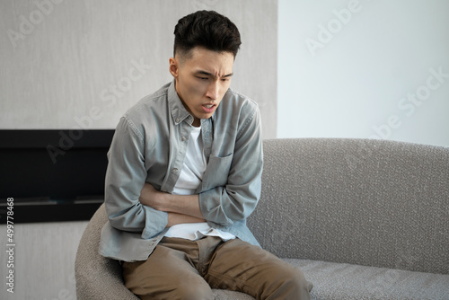 Asian man suffers from acute stomach ache sitting on sofa. Young male person in casual clothes feels abdominal pain putting hands on belly at home