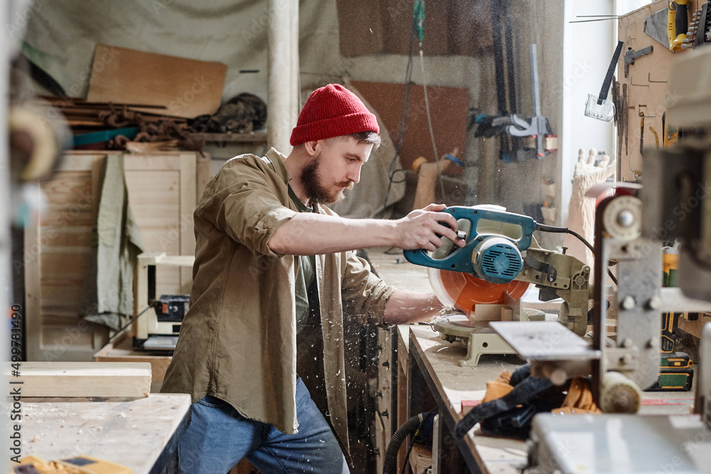 Side view of modern young Caucasian carpenter cutting wooden plank with miter saw in woodworking workshop Side view of modern young Caucasian carpenter cutting wooden plank with miter saw in woodworking workshop