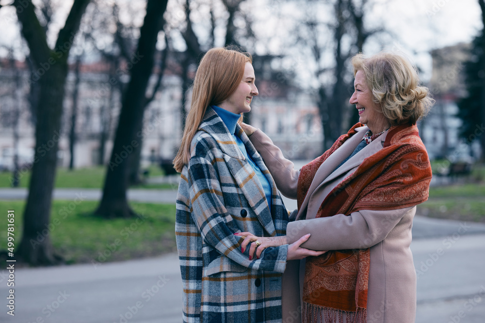 Naklejka premium A grandmother is touching her adolescent granddaughter's face with affection. They met in the park during cold weather.