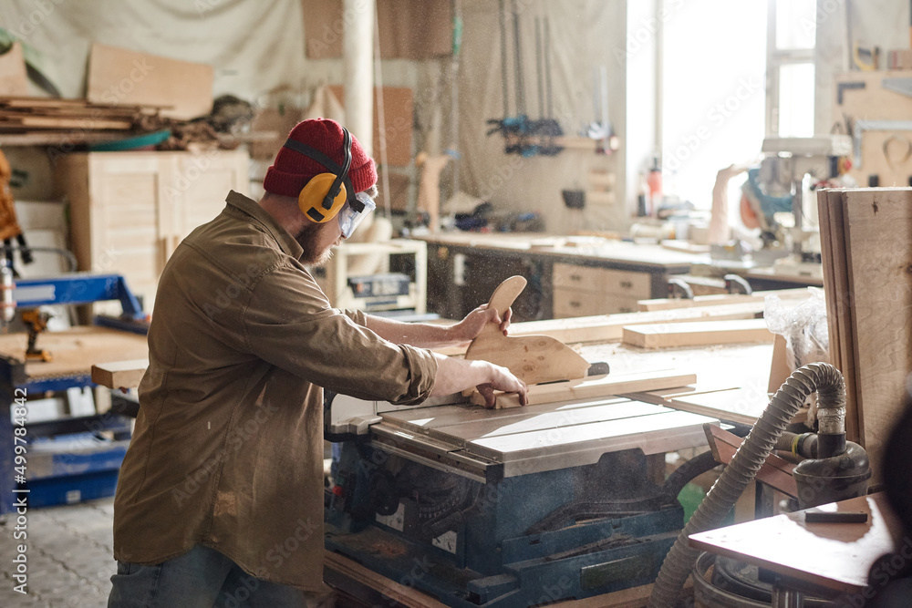 Unrecognizable carpenter wearing safety glasses and earmuffs cutting wooden board with table saw