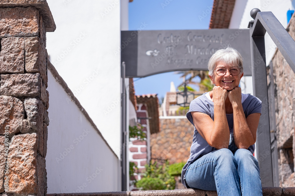 Smiling white haired senior woman sitting outdoor on staircases in a sunny day. Attractive elderly lady looking at camera enjoying relax and vacation