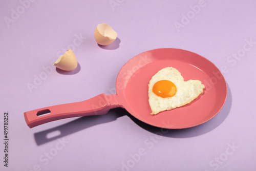 Creative idea with a pink frying pan and egg in heart shape on purple background. Minimal food and love concept. Breakfast idea for Valentine's day and romantic morning.