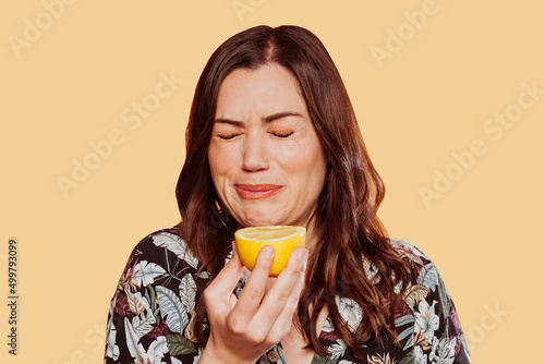 Face portrait of woman wears floral shirt, eating acid lemon doing funny face expression with eyes closed. Studio shot over yellow background. 