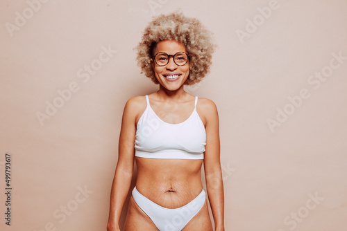Portrait of young cheerful afro latin american woman in white underwear and eyeglasses over beige background at studio. Stretch marks and cellulite on the skin. Real people, real body, natural beauty.