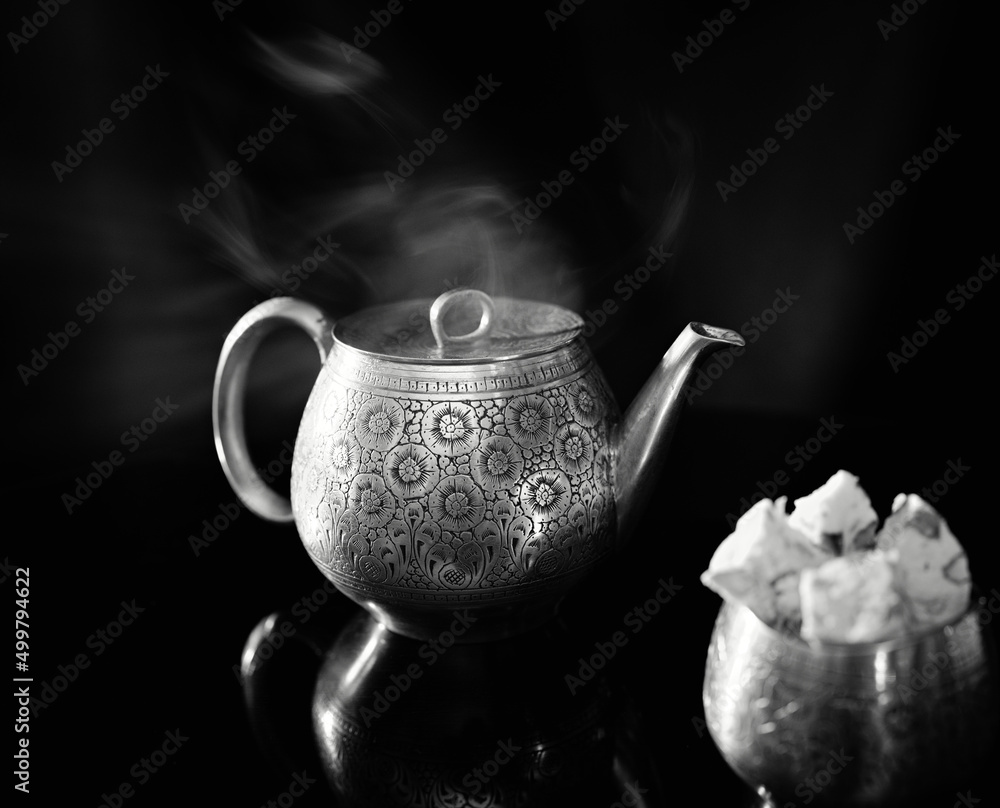 Black and white still life with a teapot and a candy bowl made of polished brass with carved oriental patterns. The brass teapot is floating, and the candy bowl is filled with onion and pistachios.