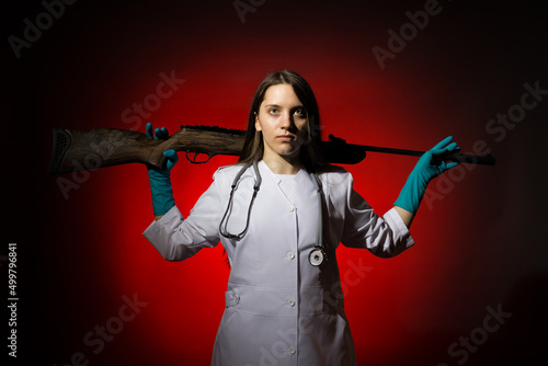 a young girl doctor in a white coat put a gun on her shoulders on a dark background © Sergey