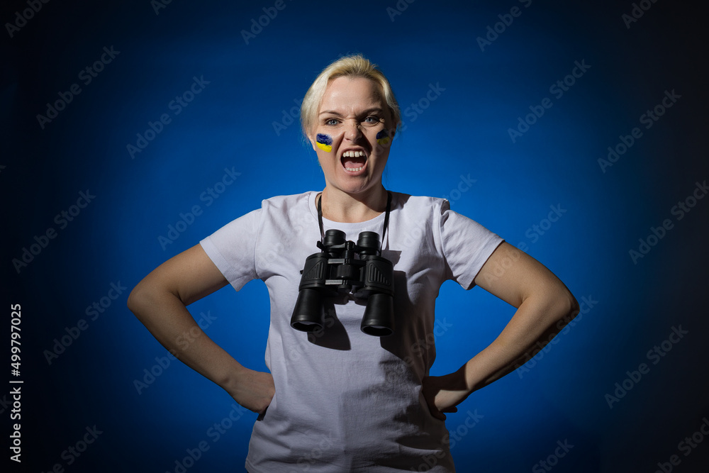 A blonde woman with a blue-yellow Ukrainian flag on her cheek with binoculars around her neck screams on a blue background. A blonde woman with a blue-yellow Ukrainian flag on her cheek with binoculars around her neck screams on a blue background.