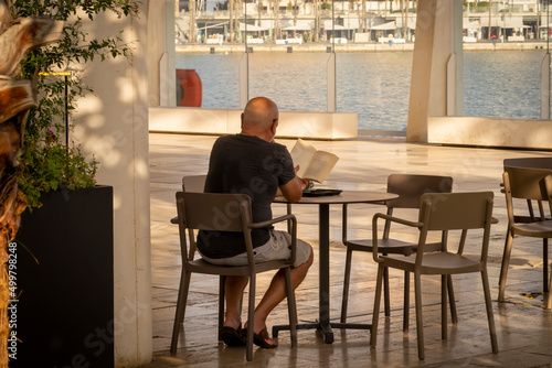 Bald man reading a book sitting on a terrace 