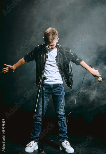 Portrait of Caucasian teenager in white t-shirt, blue jeans and leather jacket with microphone singing on dark background. Hobby and glory concept