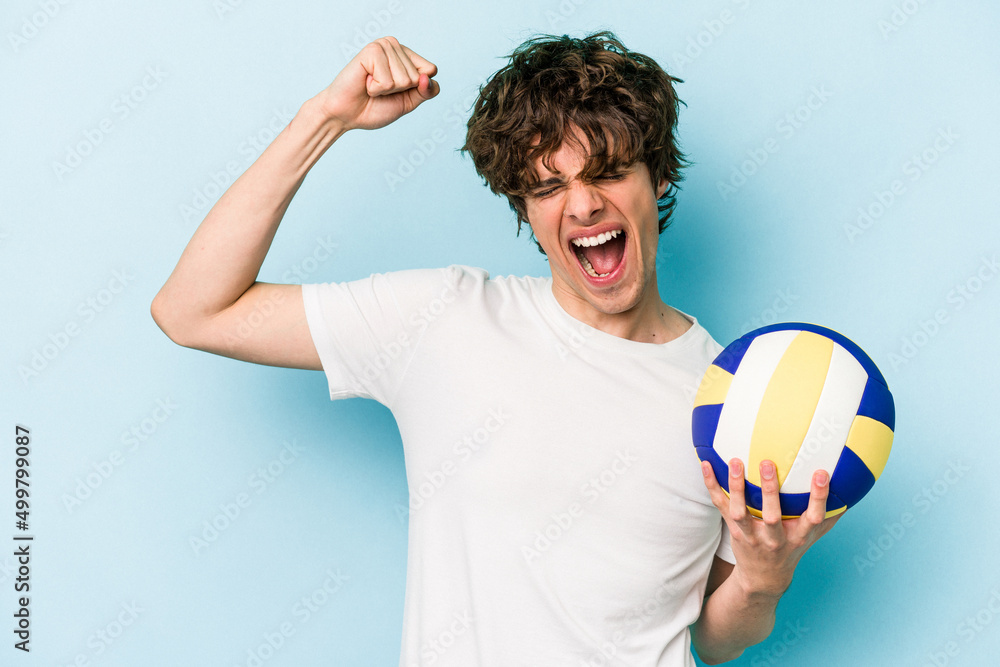 Young caucasian man playing volleyball isolated on blue background raising fist after a victory, winner concept.